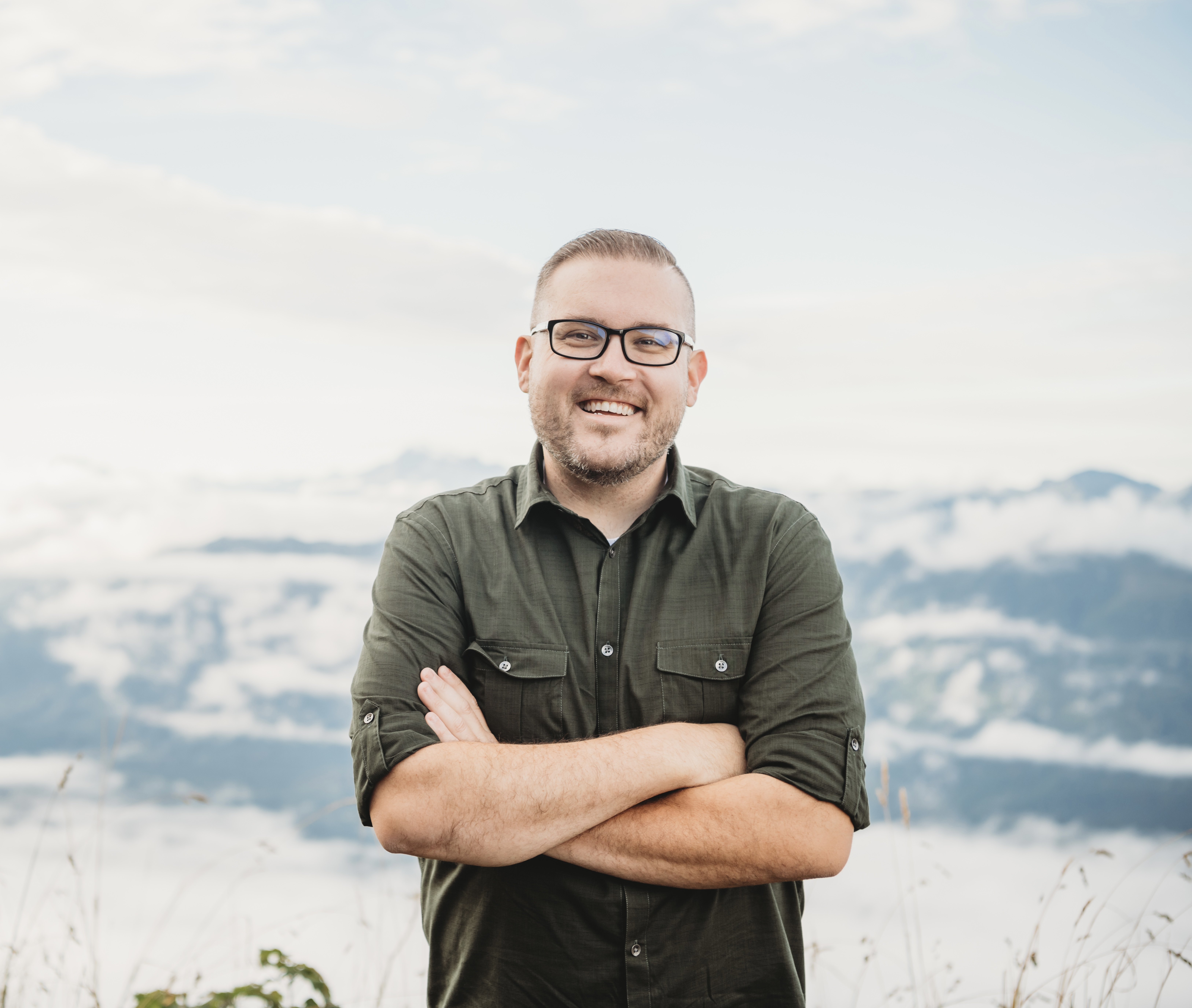 Jon smiling with arms crossed, standing above the clouds on a mountain summit