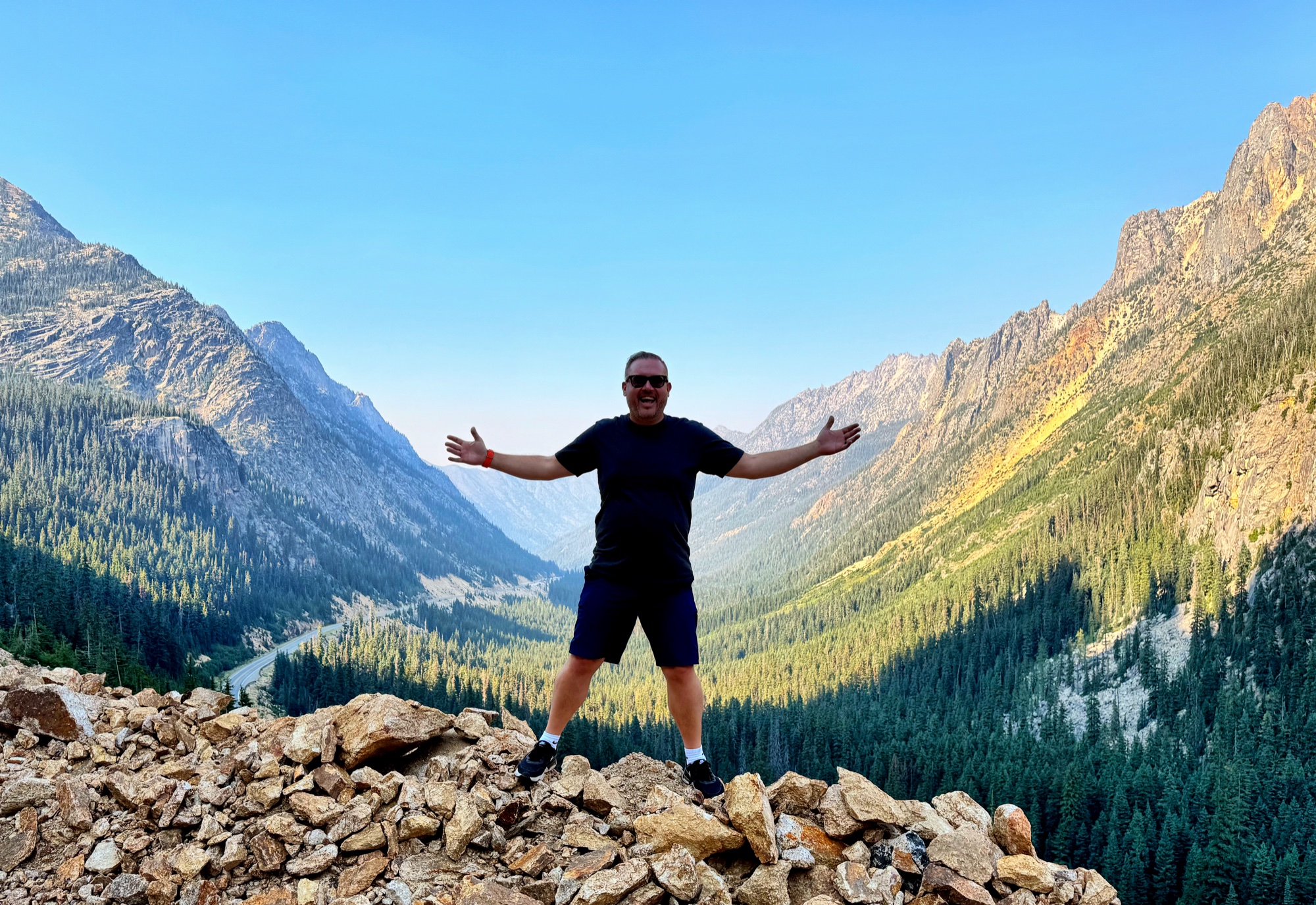 Jon standing on a rocky summit with arms wide open, epic mountain valley stretching behind him